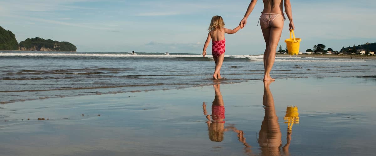 Una playa con un niño y un adulto tomados de la mano, un cubo amarillo y un reflejo sobre la arena mojada.