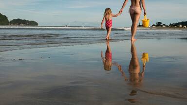 Una playa con un niño y un adulto tomados de la mano, un cubo amarillo y un reflejo sobre la arena mojada.
