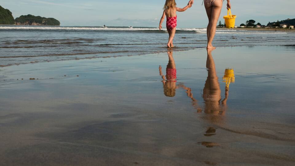 A beach with a child and an adult holding hands, a yellow bucket, and a reflection on the wet sand.