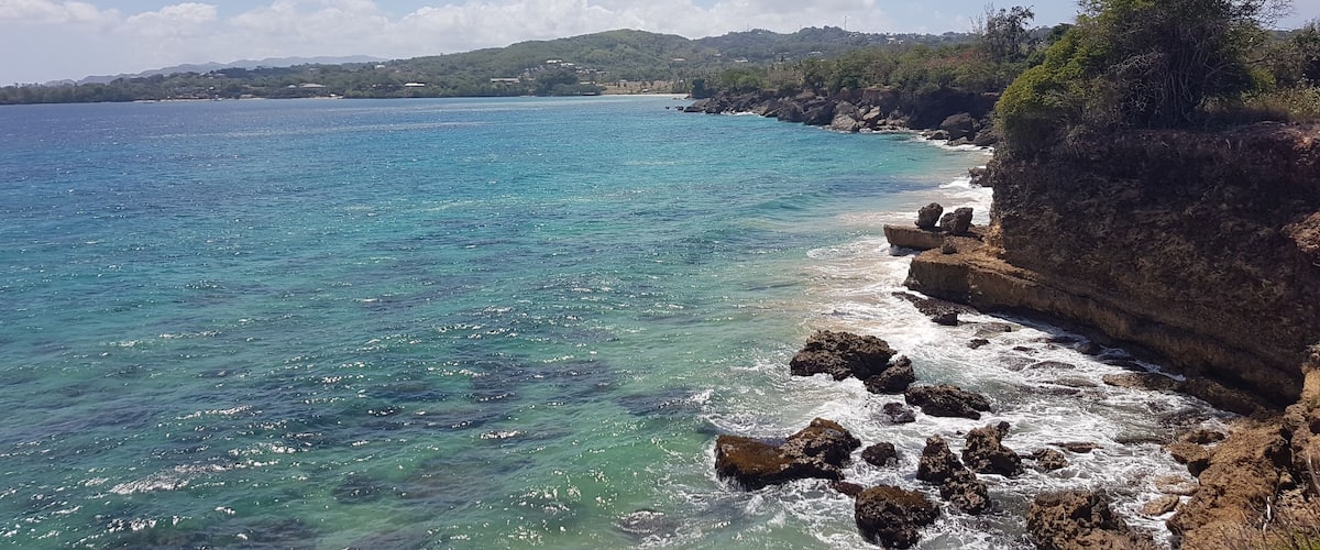 A coastal landscape with clear turquoise waters, rocky cliffs, and a distant hilly terrain.