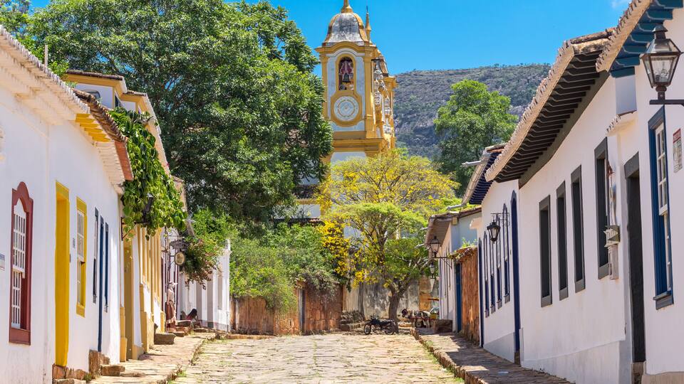 A cobblestone street lined with colorful buildings, a clock tower, and lush greenery.