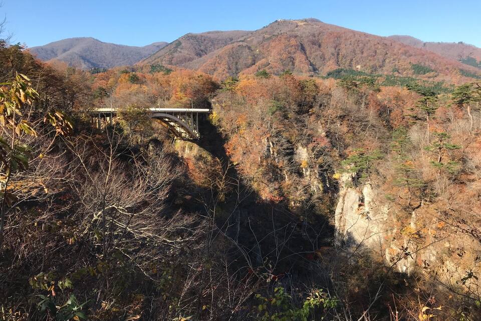 A stone bridge over a gorge with autumn foliage and mountains in the background.
