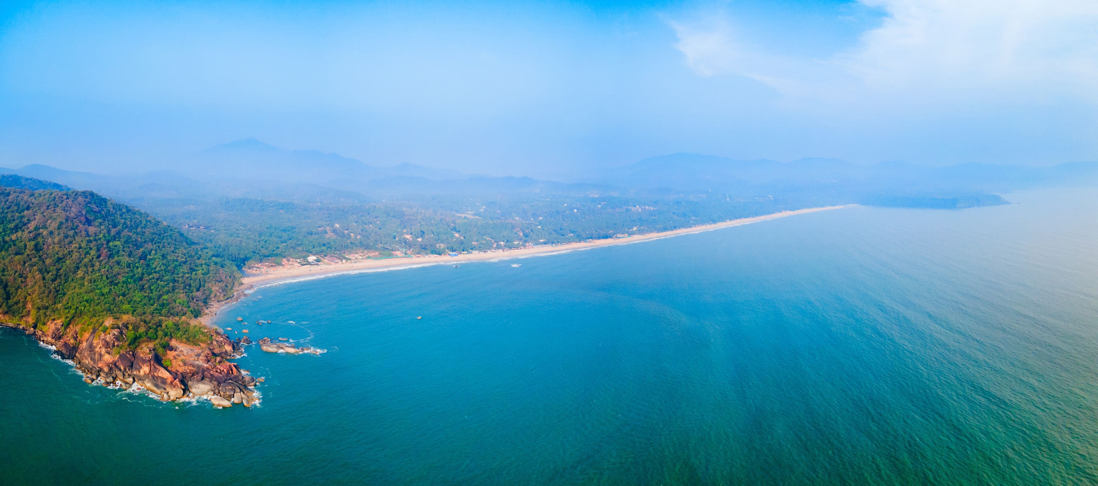 A coastal landscape with a beach, forested hills, and a clear view of the ocean.
