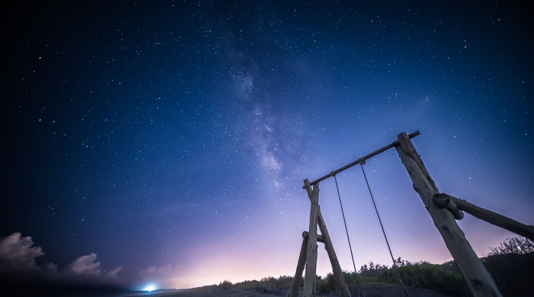 The Milky Way stretches across the night sky. A solitary wooden swing set stands on a sandy beach, inviting viewers to imagine swinging under the vast, starry sky. Zhuangwei, Yilan County, Taiwan.