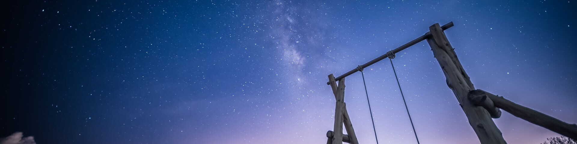 The Milky Way stretches across the night sky. A solitary wooden swing set stands on a sandy beach, inviting viewers to imagine swinging under the vast, starry sky. Zhuangwei, Yilan County, Taiwan.