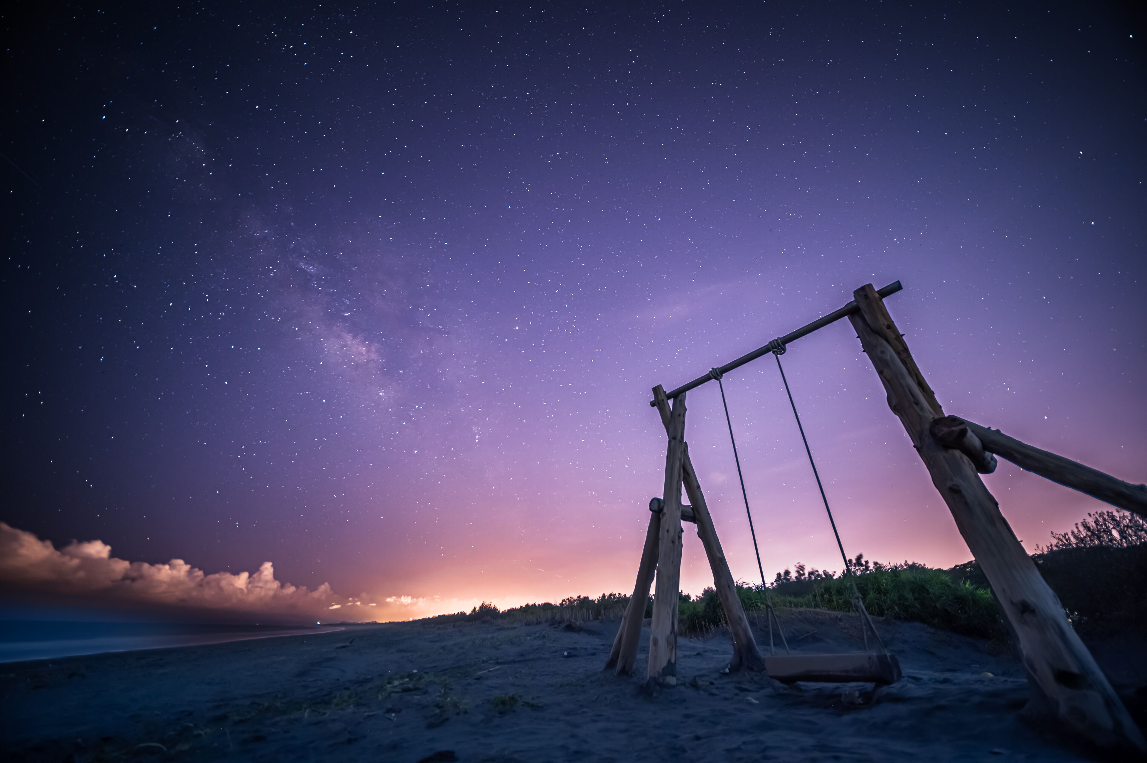The Milky Way stretches across the night sky. A solitary wooden swing set stands on a sandy beach, inviting viewers to imagine swinging under the vast, starry sky. Zhuangwei, Yilan County, Taiwan.