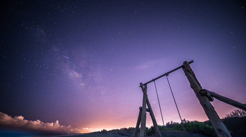The Milky Way stretches across the night sky. A solitary wooden swing set stands on a sandy beach, inviting viewers to imagine swinging under the vast, starry sky. Zhuangwei, Yilan County, Taiwan.