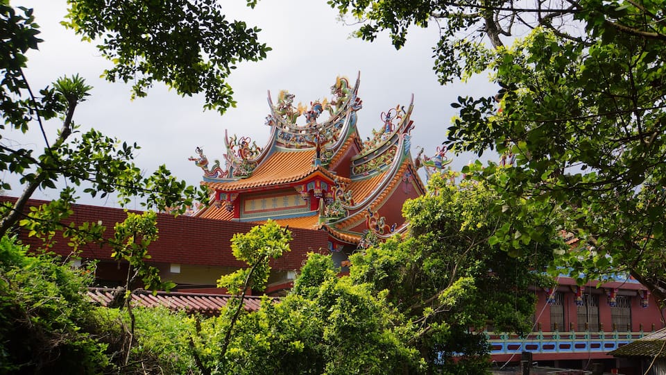 A traditional temple with ornate architecture and a red roof, surrounded by lush green trees.