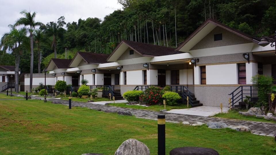 A row of modern buildings with sloped roofs, surrounded by lush greenery and palm trees.