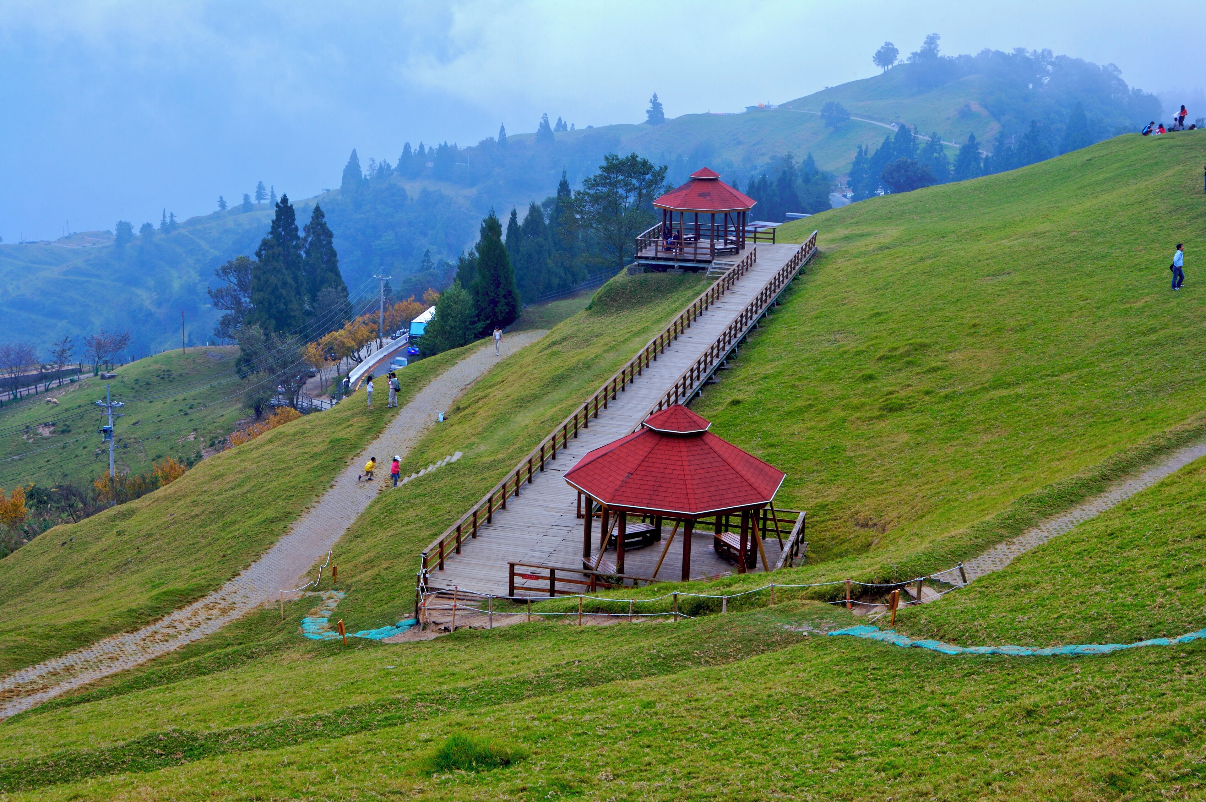 A wooden observation deck with a red roof on a grassy hill, overlooking a valley with trees and a distant mountain range.