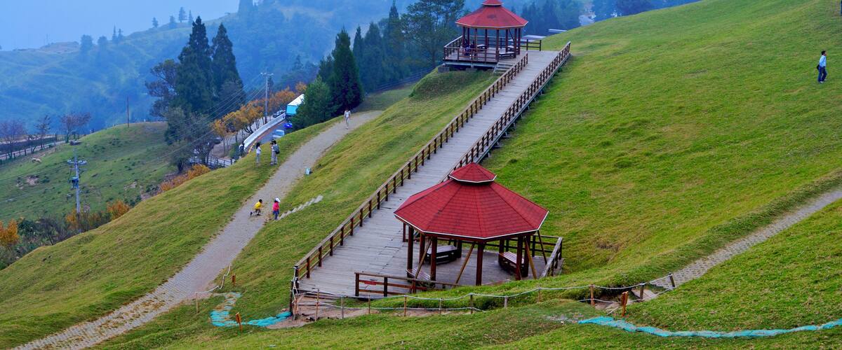 A wooden observation deck with a red roof on a grassy hill, overlooking a valley with trees and a distant mountain range.
