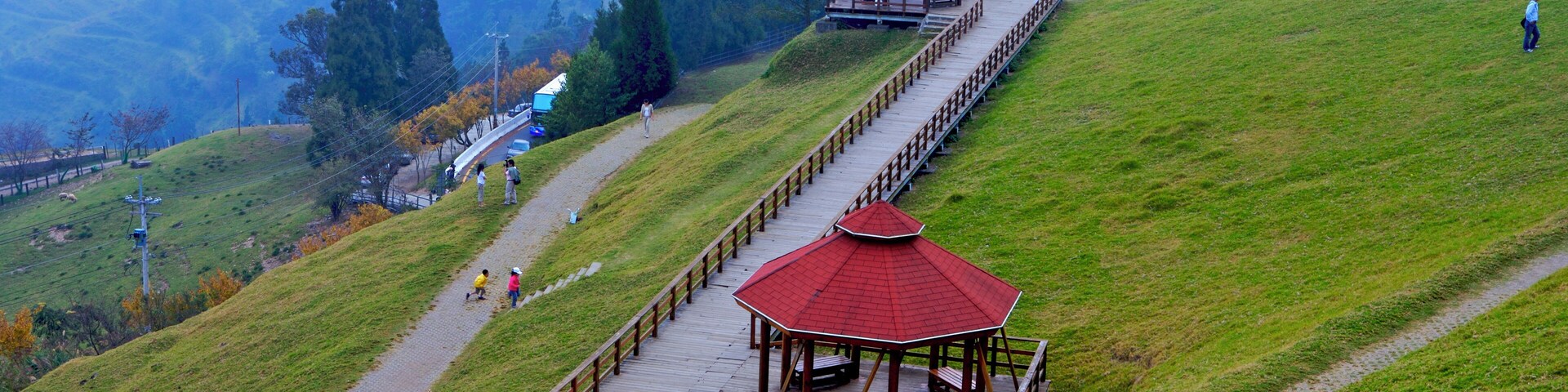 A wooden observation deck with a red roof on a grassy hill, overlooking a valley with trees and a distant mountain range.