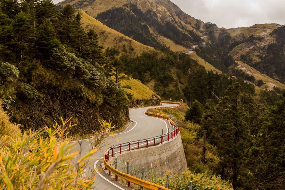 A winding mountain road with guardrails, surrounded by dense forests and a prominent mountain in the background.