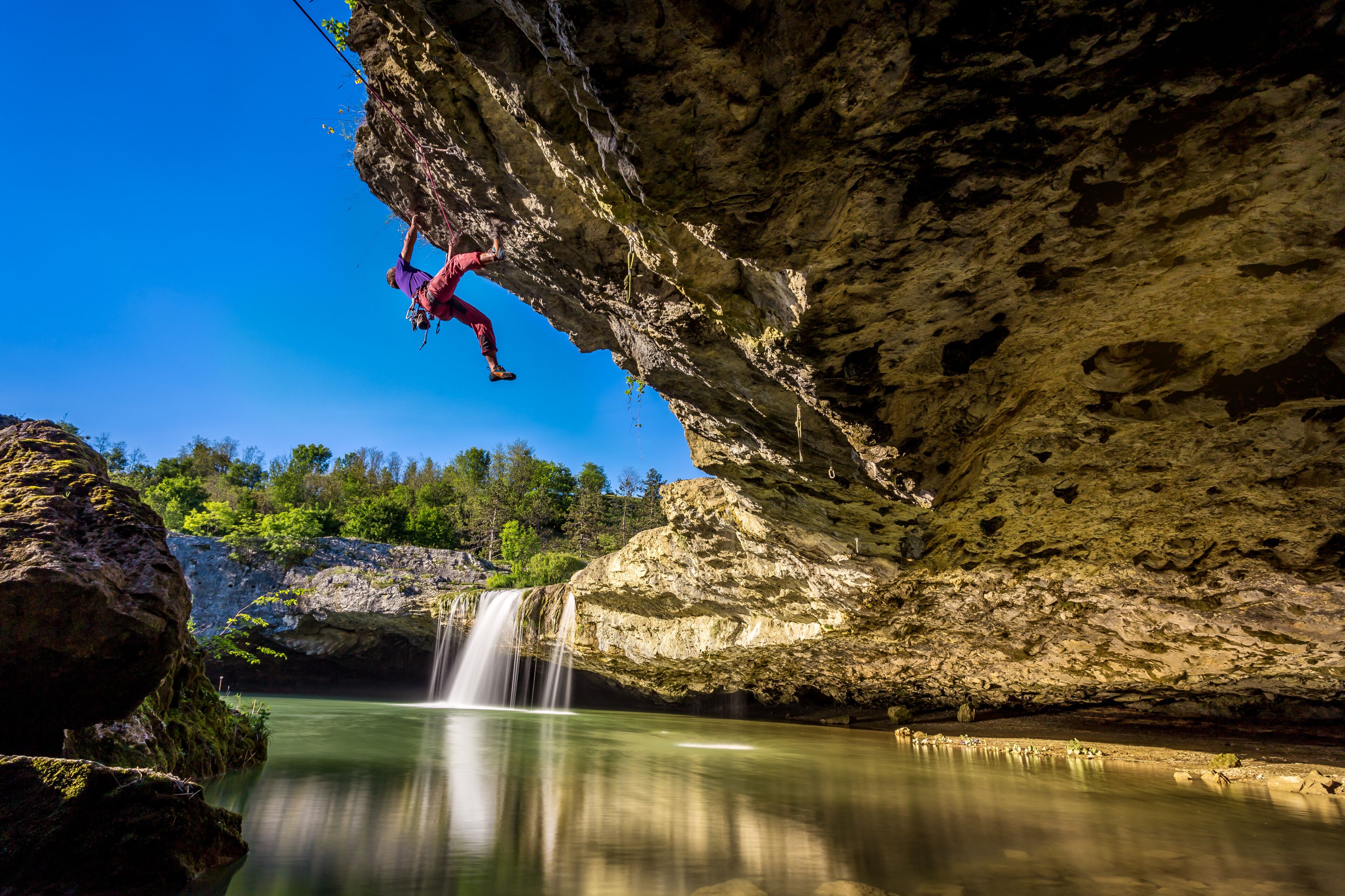 View of a climber suspended beneath a rocky overhang as a waterfall cascades into the still, reflective waters below, Pazin, Istria County, Croatia.