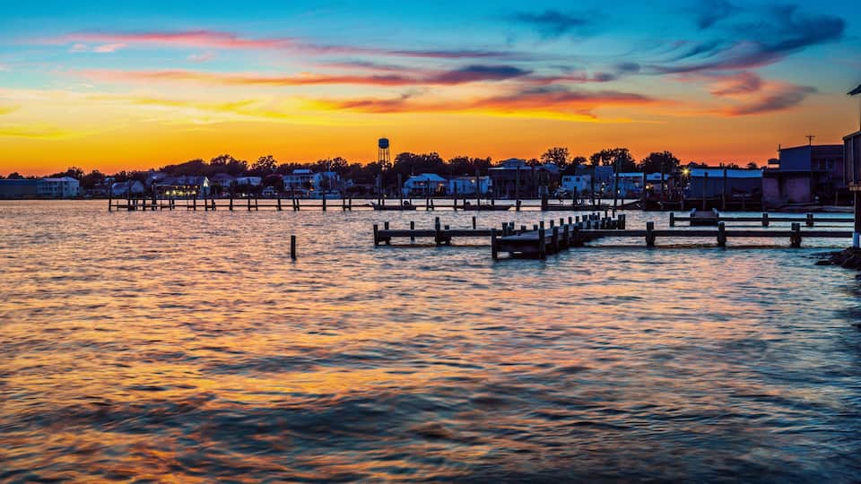 A waterfront at sunset with a pier extending into the water, buildings along the shore, and a water tower in the background.