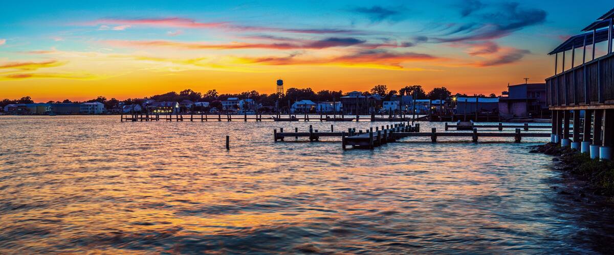 A waterfront at sunset with a pier extending into the water, buildings along the shore, and a water tower in the background.