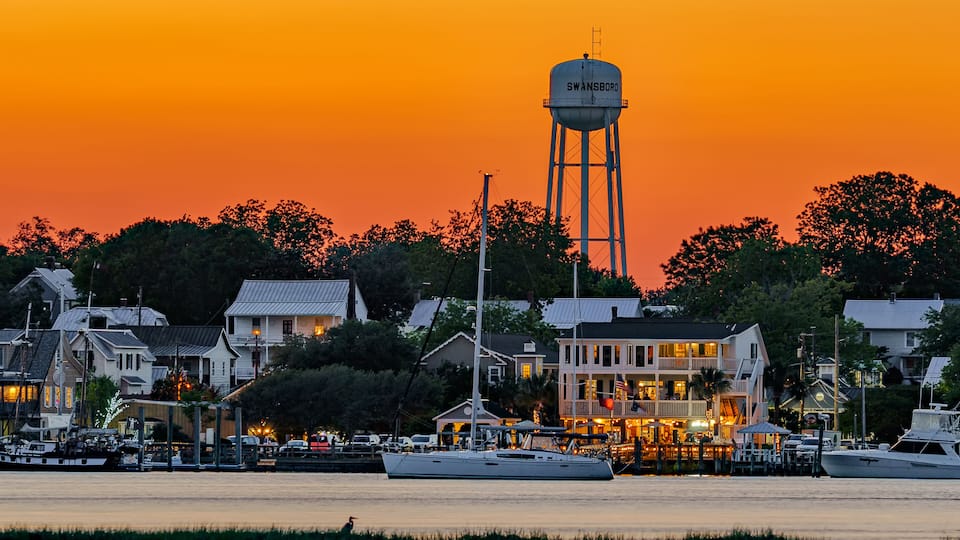 A waterfront town at sunset with a prominent water tower, docked boats, and residential buildings.