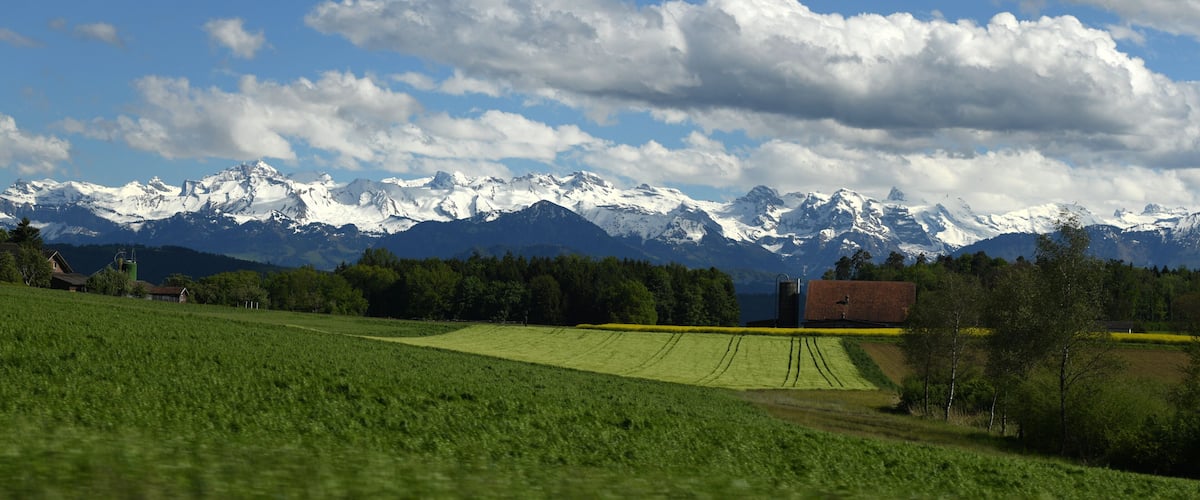A green field, a red barn, and snow-capped mountains under a blue sky.