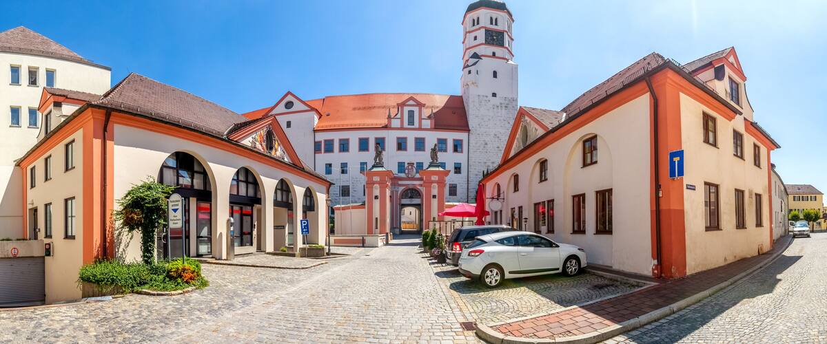 A cobblestone street with historic buildings, a prominent tower, and a white car parked on the side.