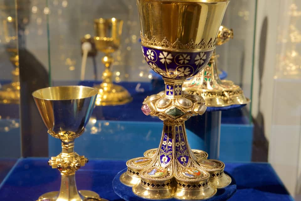 A display of ornate religious artifacts, including a golden chalice and a decorated paten, on a blue velvet cloth.