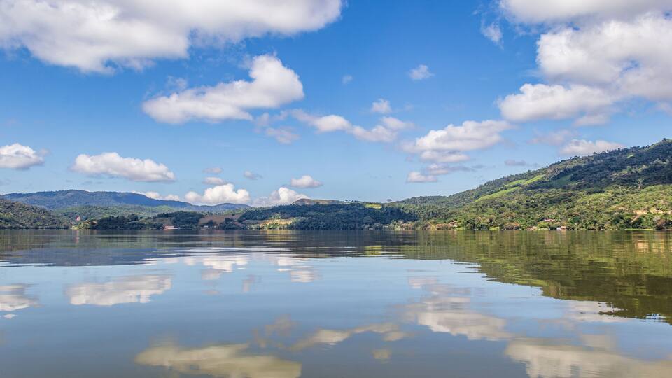 A calm lake with reflections of clouds, surrounded by green hills and a clear blue sky.