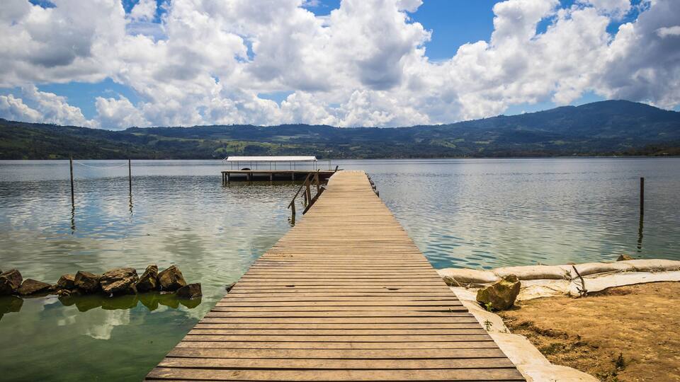 A wooden pier extending into a calm lake with a white canopy at the end, surrounded by mountains and a partly cloudy sky.