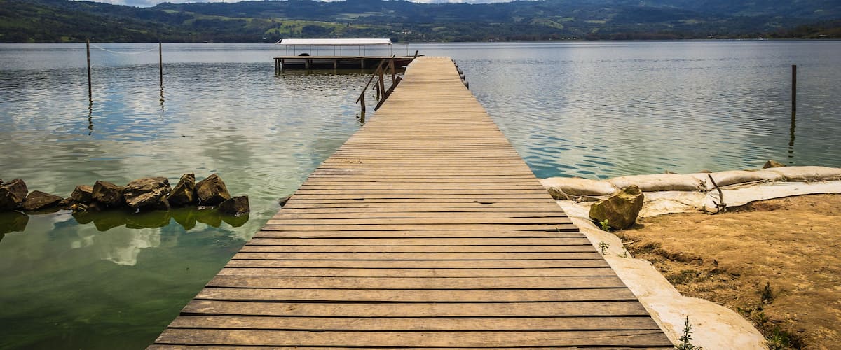 A wooden pier extending into a calm lake with a white canopy at the end, surrounded by mountains and a partly cloudy sky.