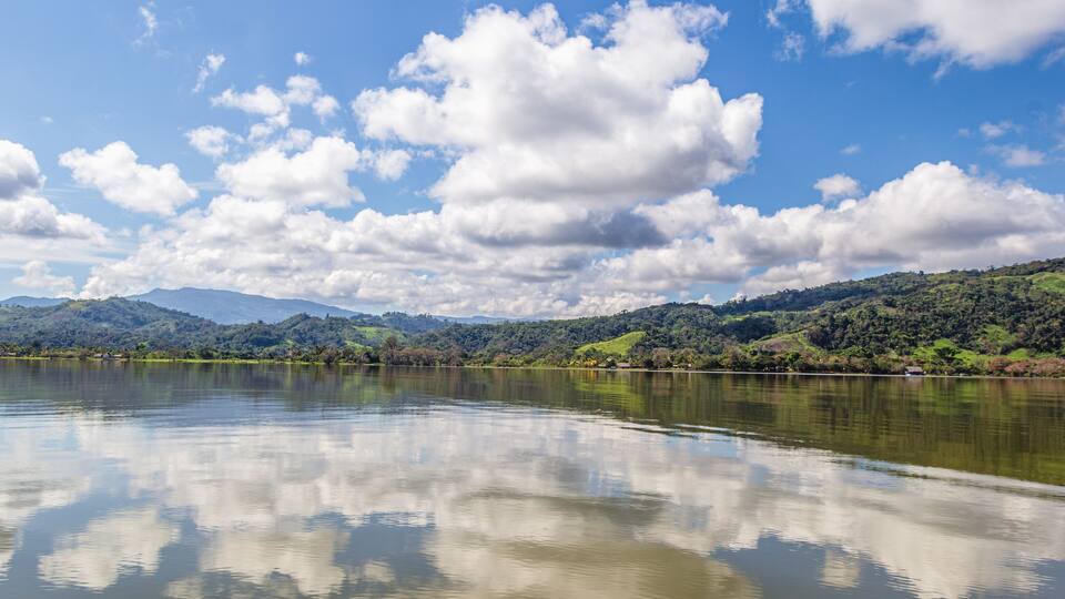 A calm lake with reflections of clouds, surrounded by green hills and mountains under a clear blue sky.