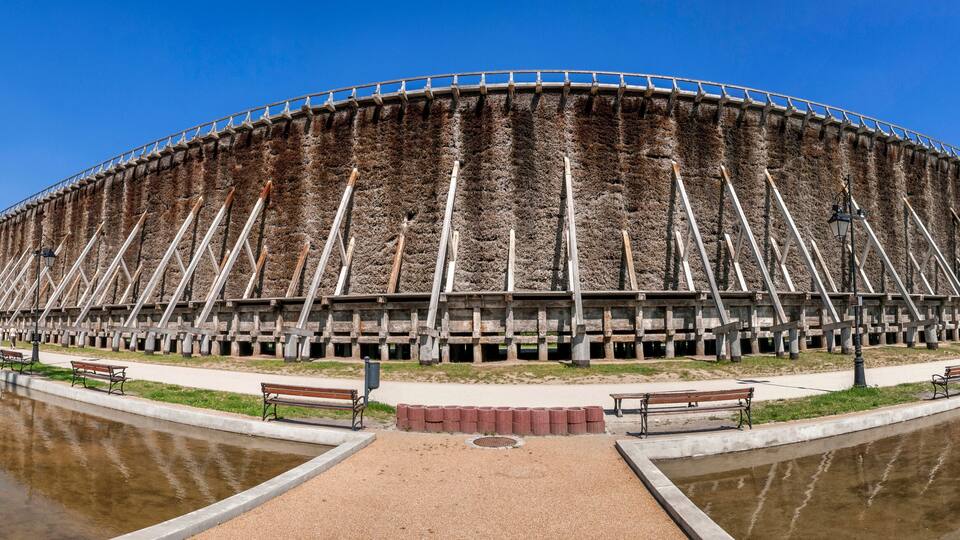 A large, circular building with a unique, layered exterior, surrounded by a body of water and benches along the walkway.