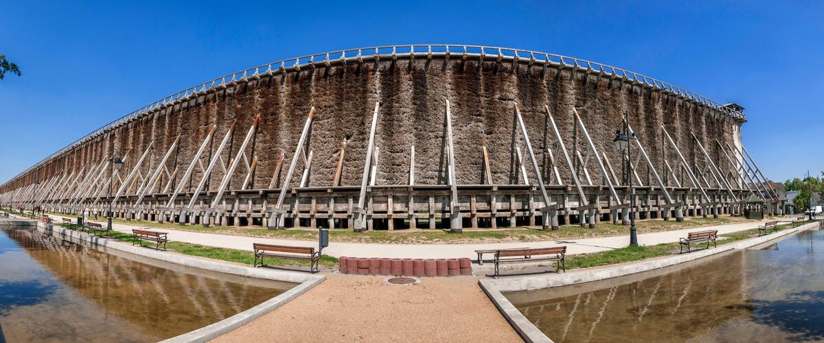 A large, circular building with a unique, layered exterior, surrounded by a body of water and benches along the walkway.