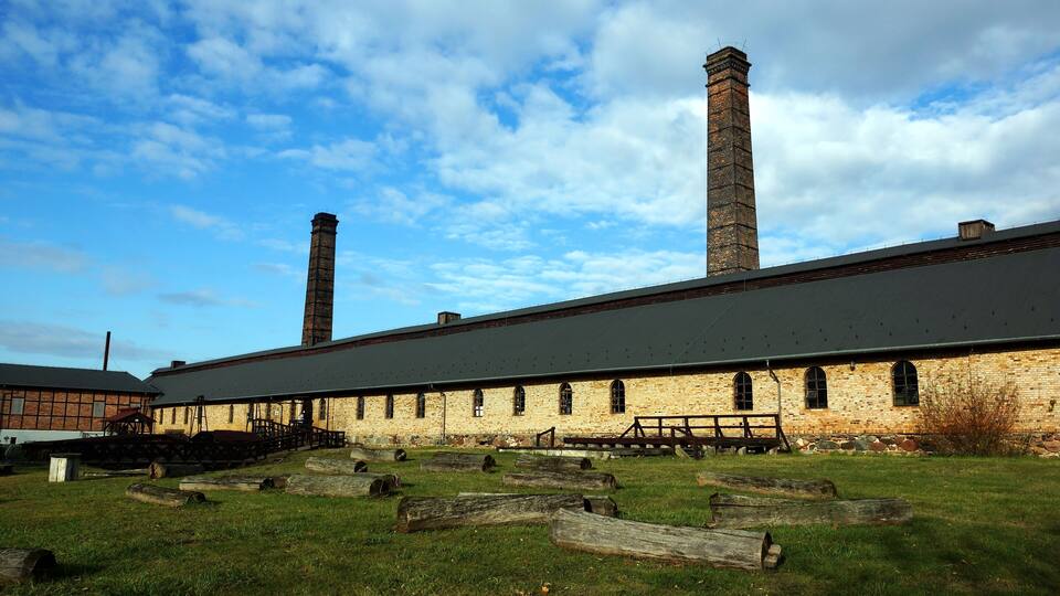 An industrial building with two chimneys, a grassy field, and wooden logs.