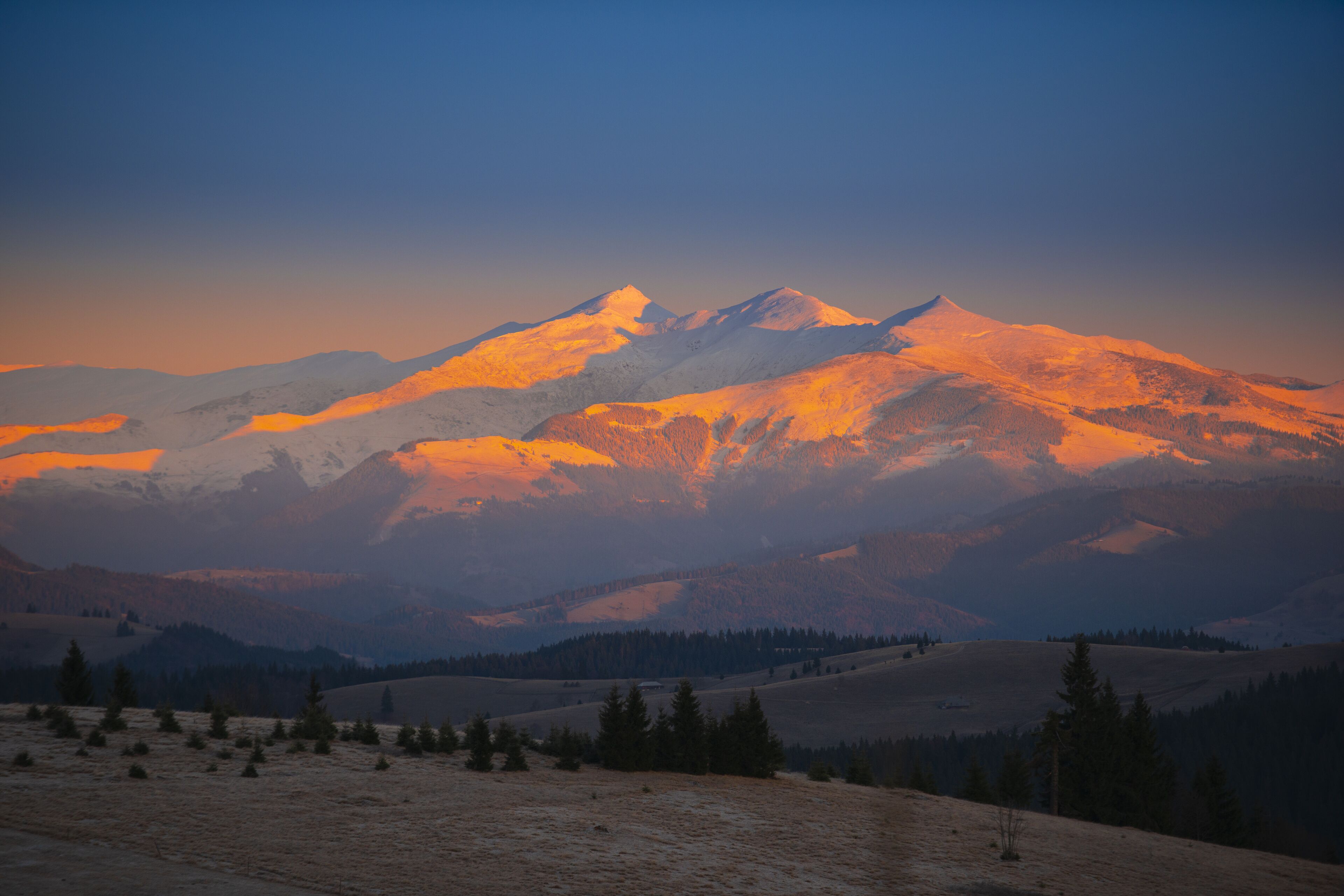 A mountain range at sunset with snow-capped peaks and a forested valley.