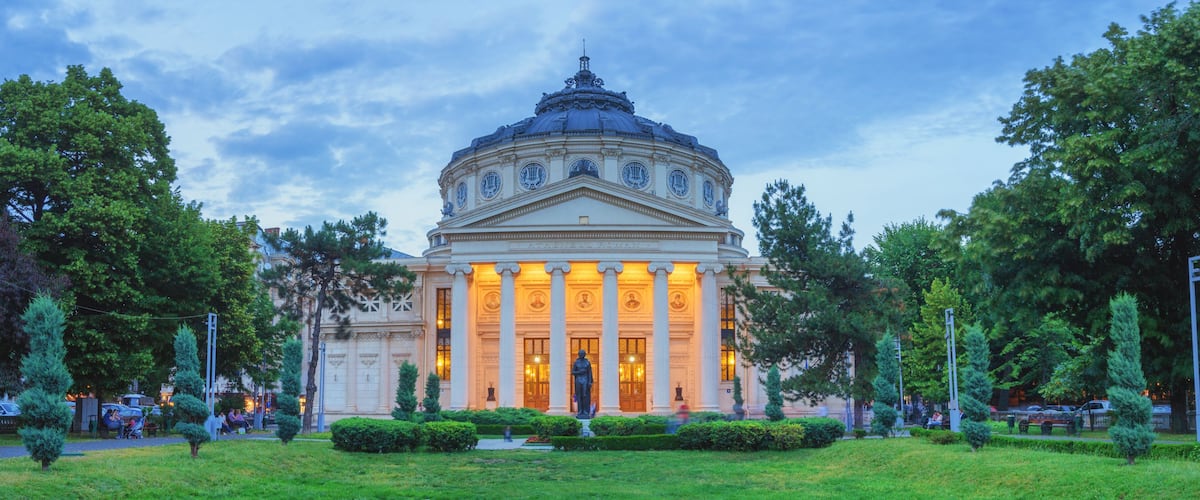 A grand neoclassical building with a dome and columns, surrounded by lush greenery and trees.