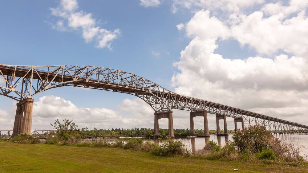 A large steel bridge spanning a river with grassy banks and trees in the background.