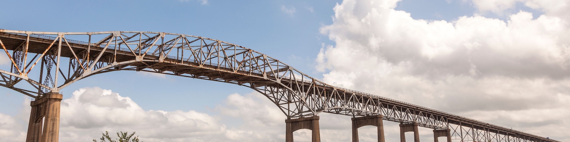 Un puente de acero grande que cruza un río con orillas cubiertas de hierba y árboles al fondo.