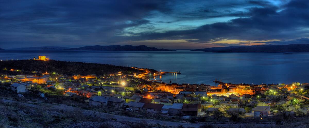 A coastal town at night with illuminated buildings and a calm sea under a cloudy sky.