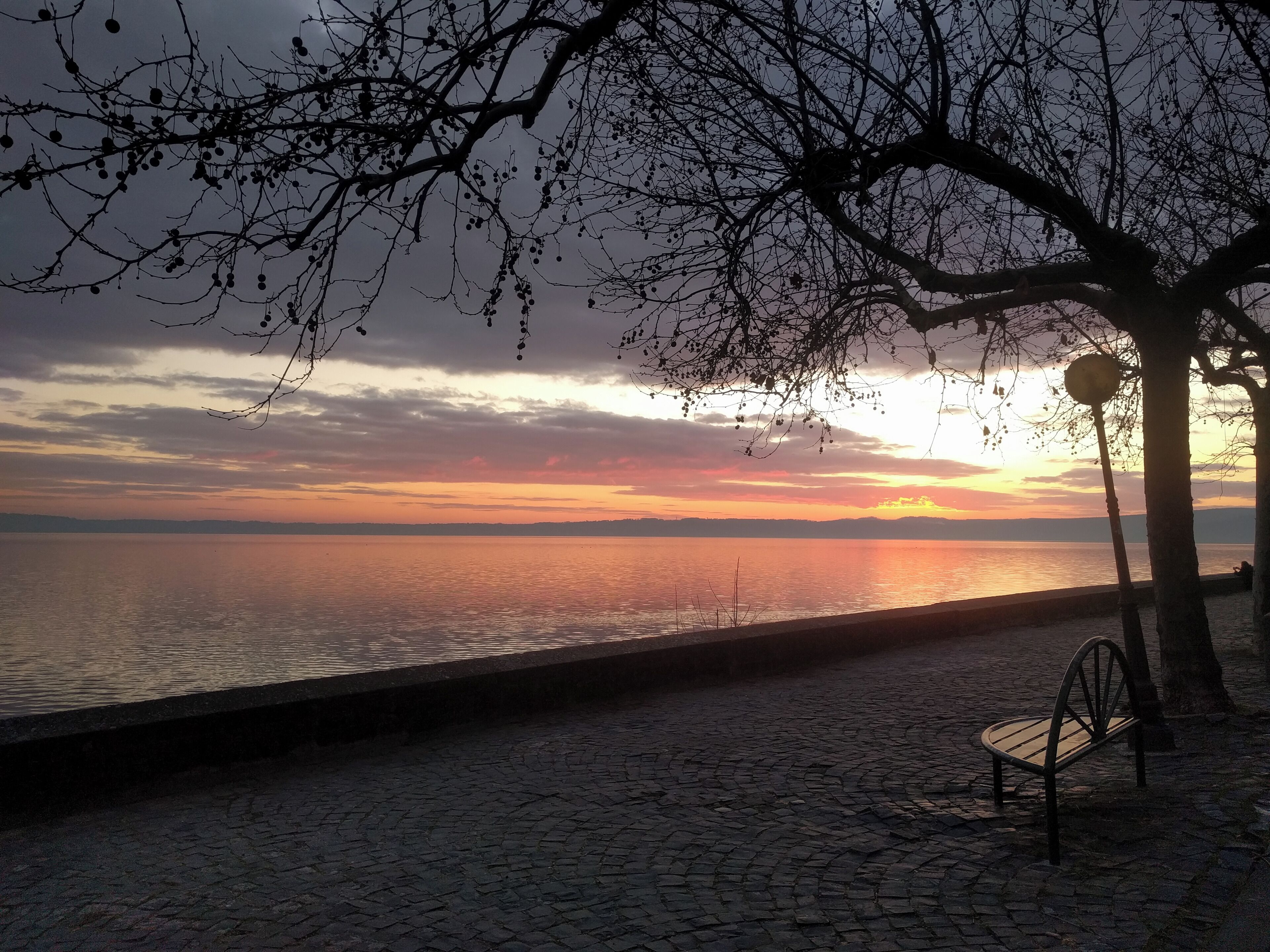 A sunset over a calm body of water with a cobblestone path and a bench.