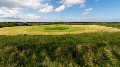 A circular field with a central green area, surrounded by a grassy perimeter, under a partly cloudy sky.