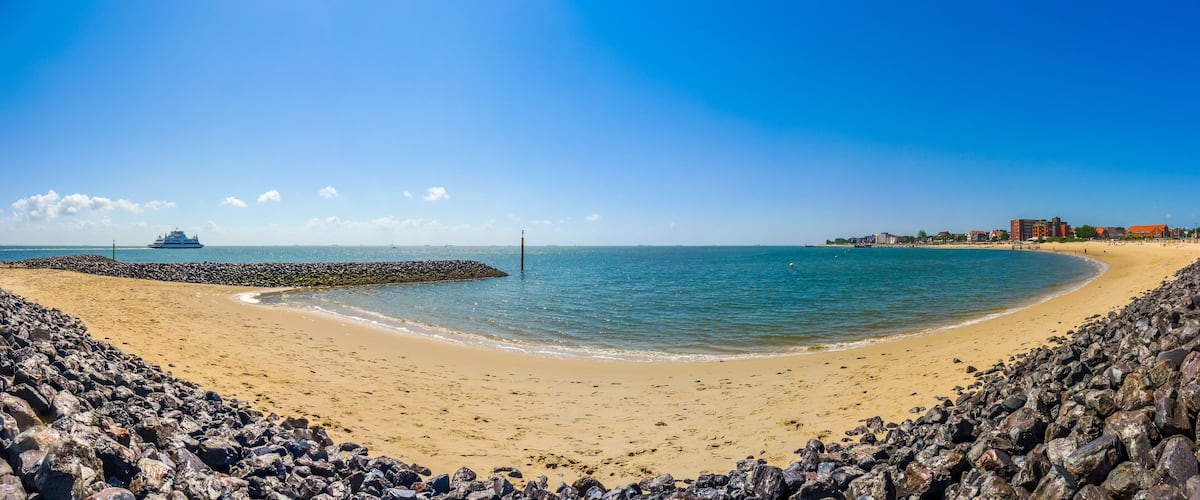 A sandy beach with a rocky breakwater, calm sea, and buildings in the background.