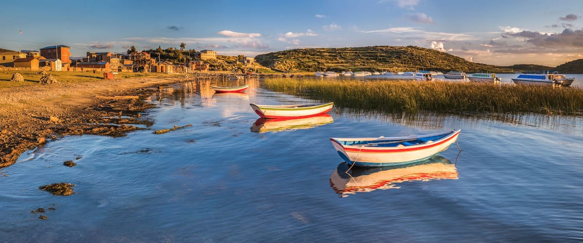 A calm lake with boats, a shoreline with buildings, and a hill in the background.