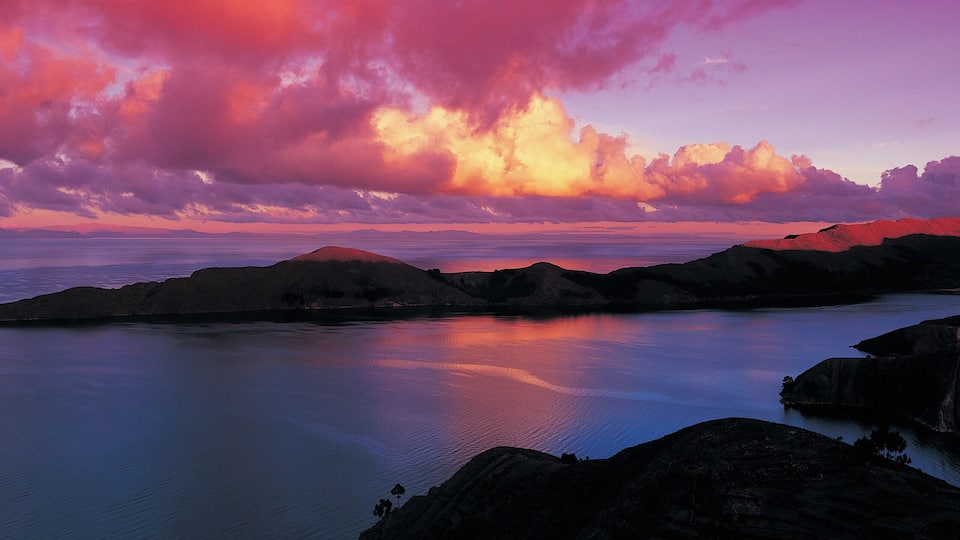 A coastal landscape with a calm sea, rocky formations, and a dramatic sunset sky.