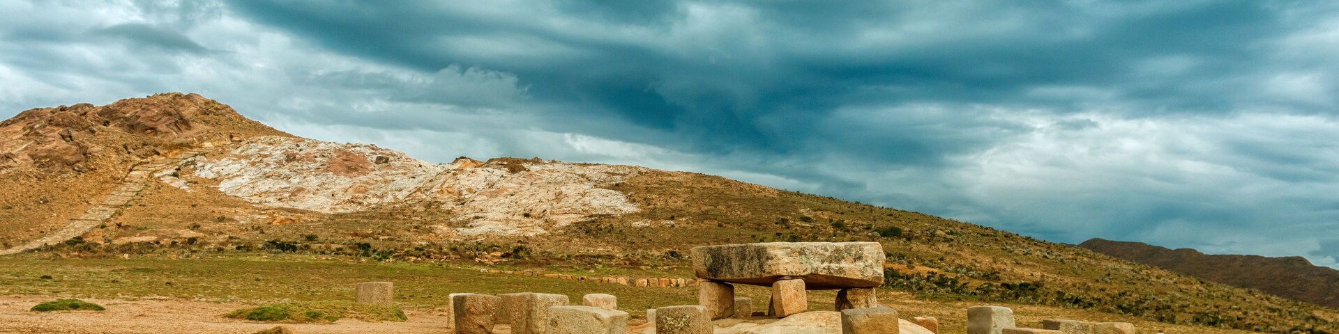 Una estructura de piedra con un pilar central y bloques circundantes, frente a un paisaje montañoso bajo un cielo nublado.
