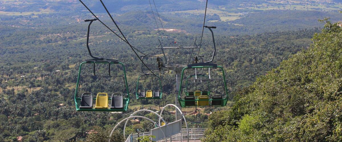 A cable car system with green chairs and yellow seats, suspended over a lush green landscape with distant city buildings.