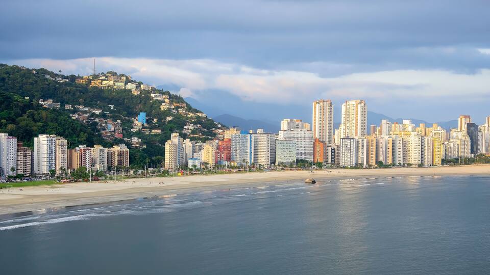 A coastal cityscape with high-rise buildings, a beach, and a hill with greenery.