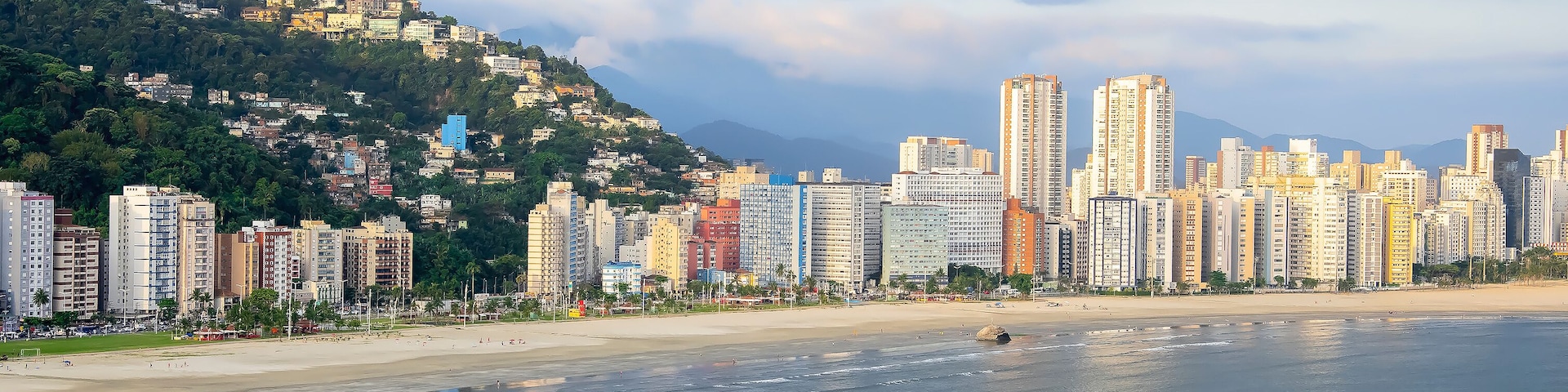 A coastal cityscape with high-rise buildings, a beach, and a hill with greenery.