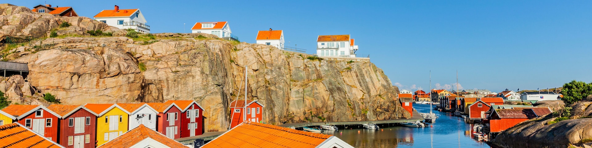 Uma vila costeira com casas de madeira coloridas, um penhasco rochoso e águas calmas.