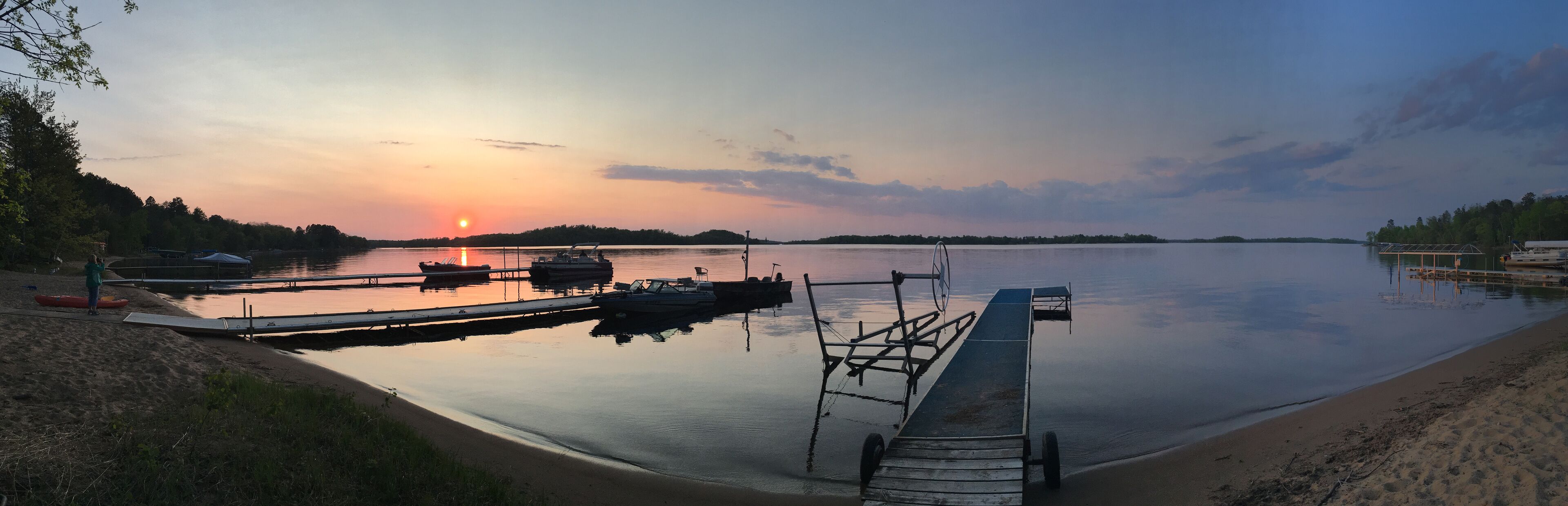 A calm lake at sunset with boats docked, a dock, and a sandy shore.