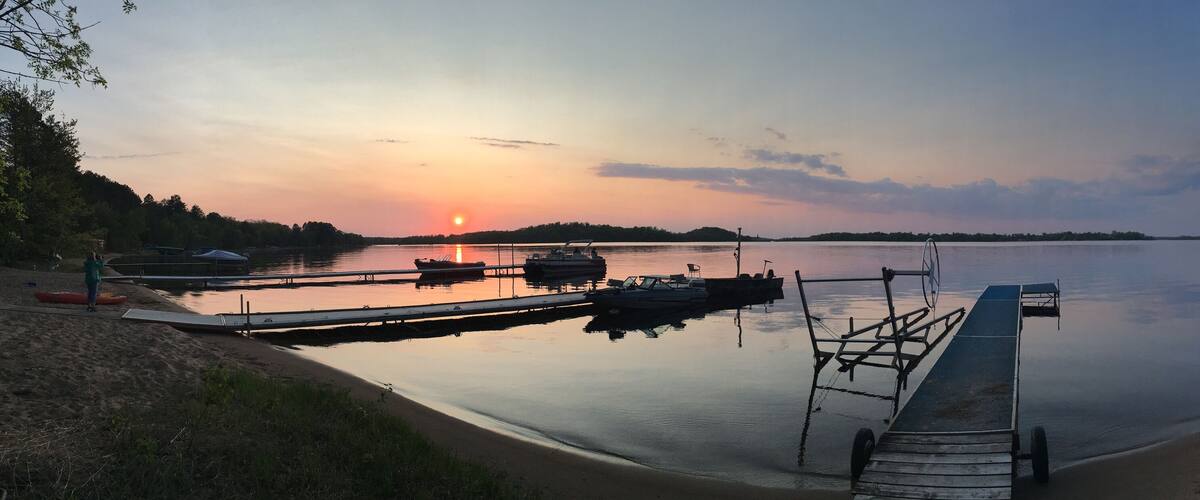 A calm lake at sunset with boats docked, a dock, and a sandy shore.