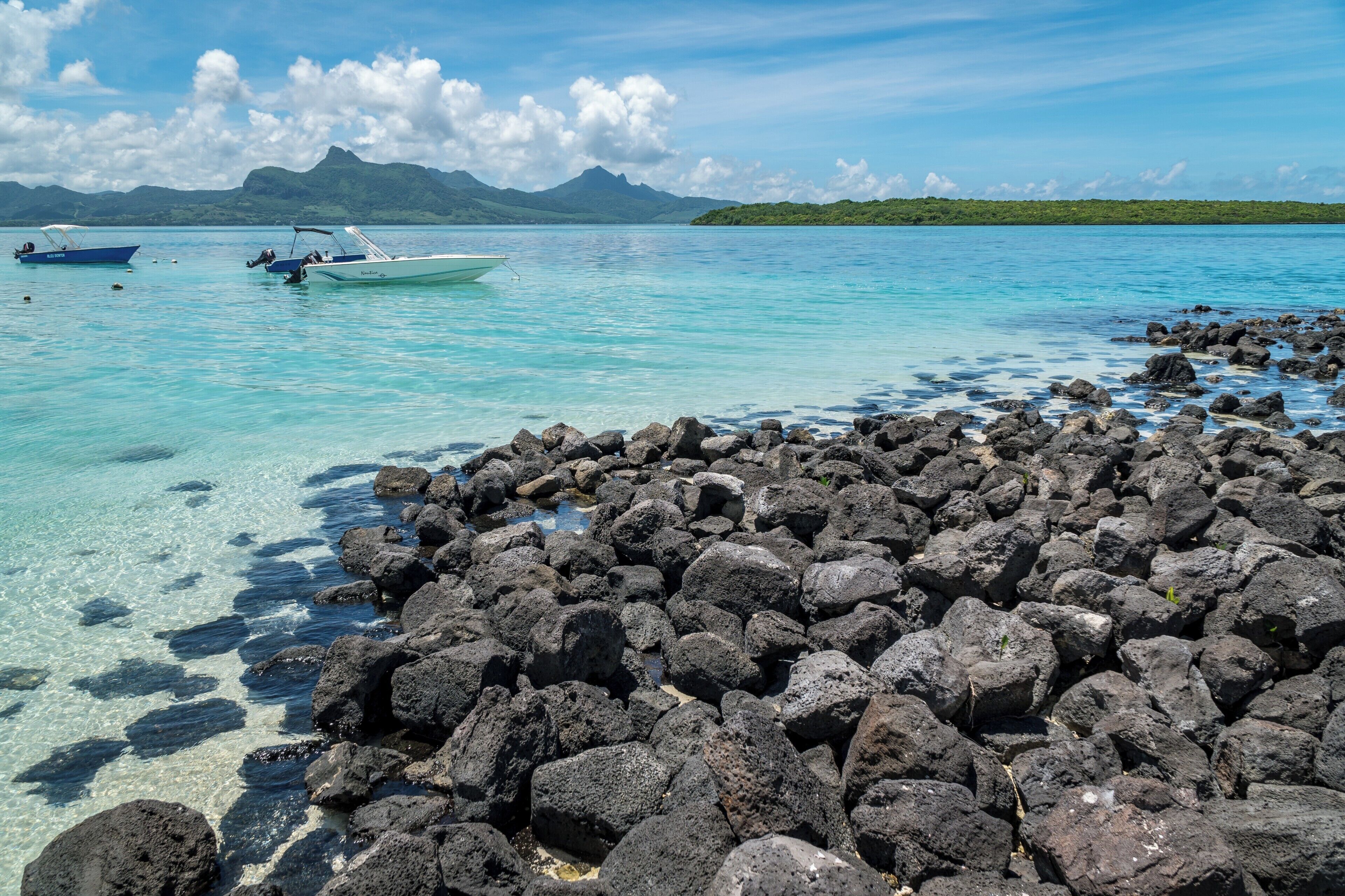 A rocky shoreline with clear turquoise water, two boats, and distant mountains.
