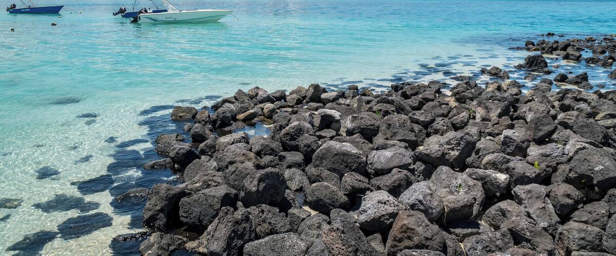 A rocky shoreline with clear turquoise water, two boats, and distant mountains.